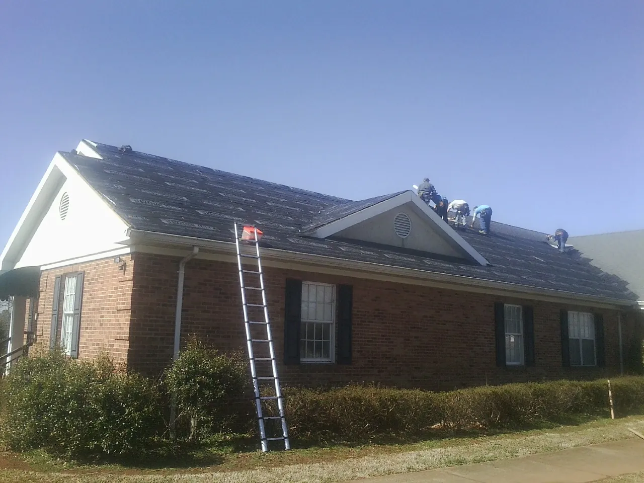 Crew tearing shingles off a brick ranch roof with a ladder set — Metal Roof Repair work by our crew in Colts Neck