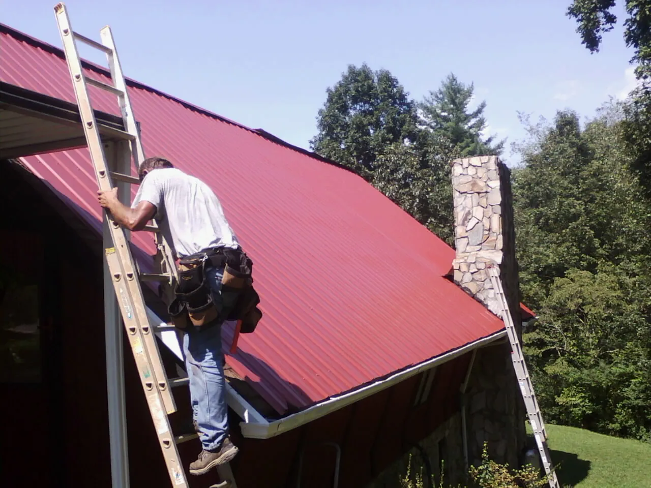 Roofer on a ladder beside a red metal roof and stone chimney — Soffit & Fascia Repair by Roofing Brothers in Colts Neck