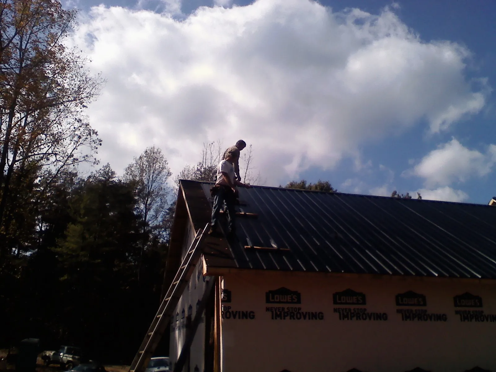 Roofer fastening dark metal panels high against a cloudy sky, from a Roof Inspection job in Colts Neck